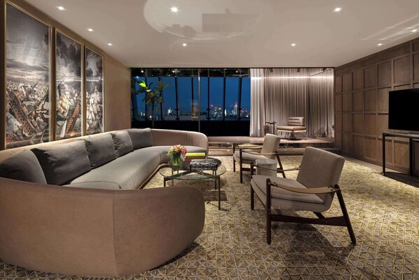 Una sala de estar moderna con un sofá modular grande, una mesa de centro de vidrio y un sillón c ómodo. Un televisor de pantalla plana está montado en la pared, y la habitación cuenta con un ventanal con vistas a la ciudad.