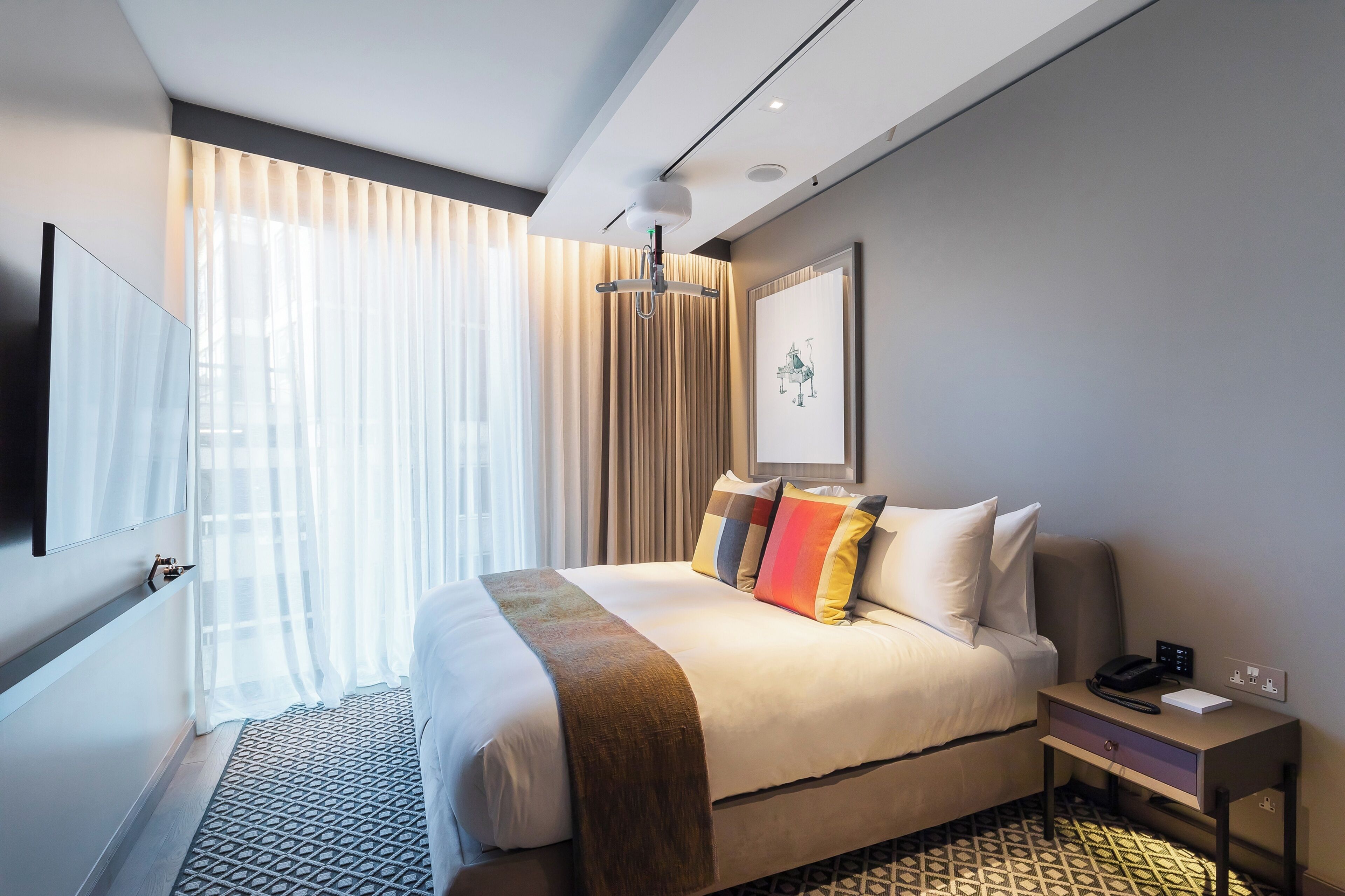 Camera d'hotel moderna con un letto grande, un comodino con telefono e lampada, una TV a parete e una finestra con tende leggere.
