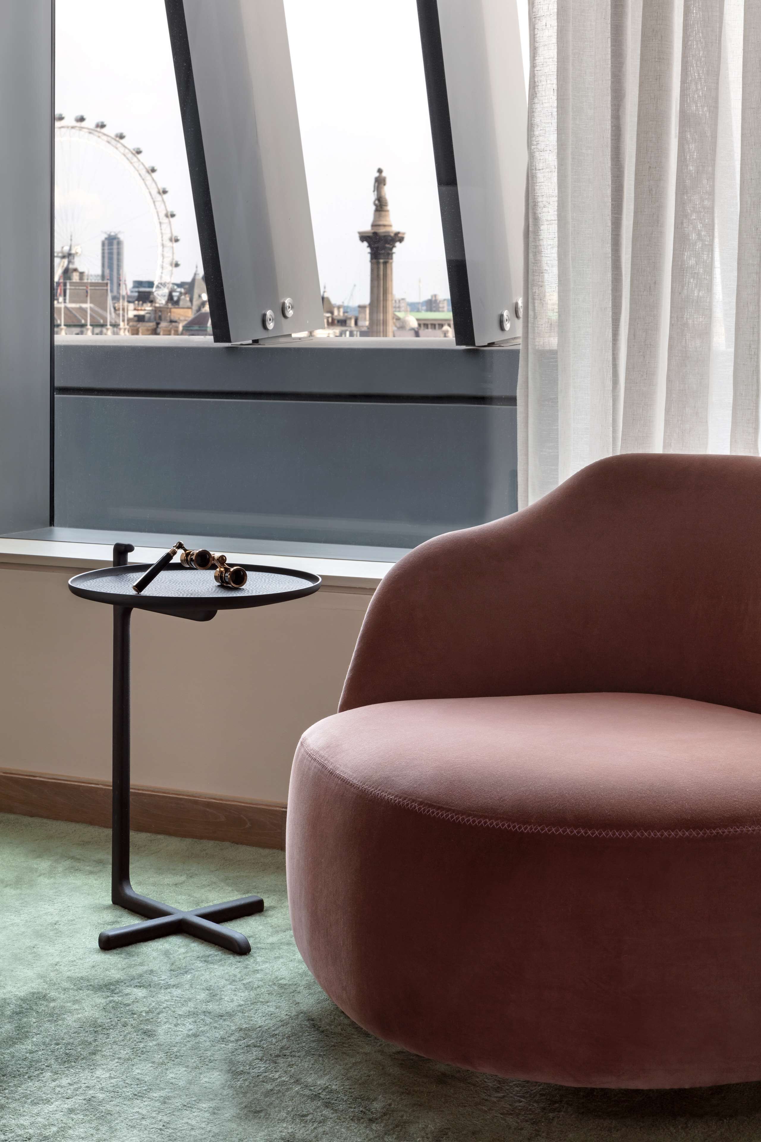 A room with a pink chair, a small round table with a tray, and a view of a Ferris wheel and tower through large windows.