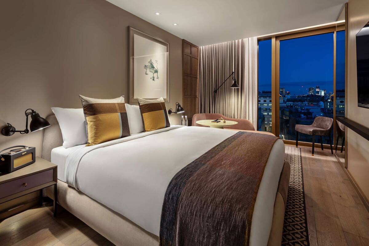A modern hotel room with a large bed, bedside tables, a small round table, a chair, and a view of the city at night.