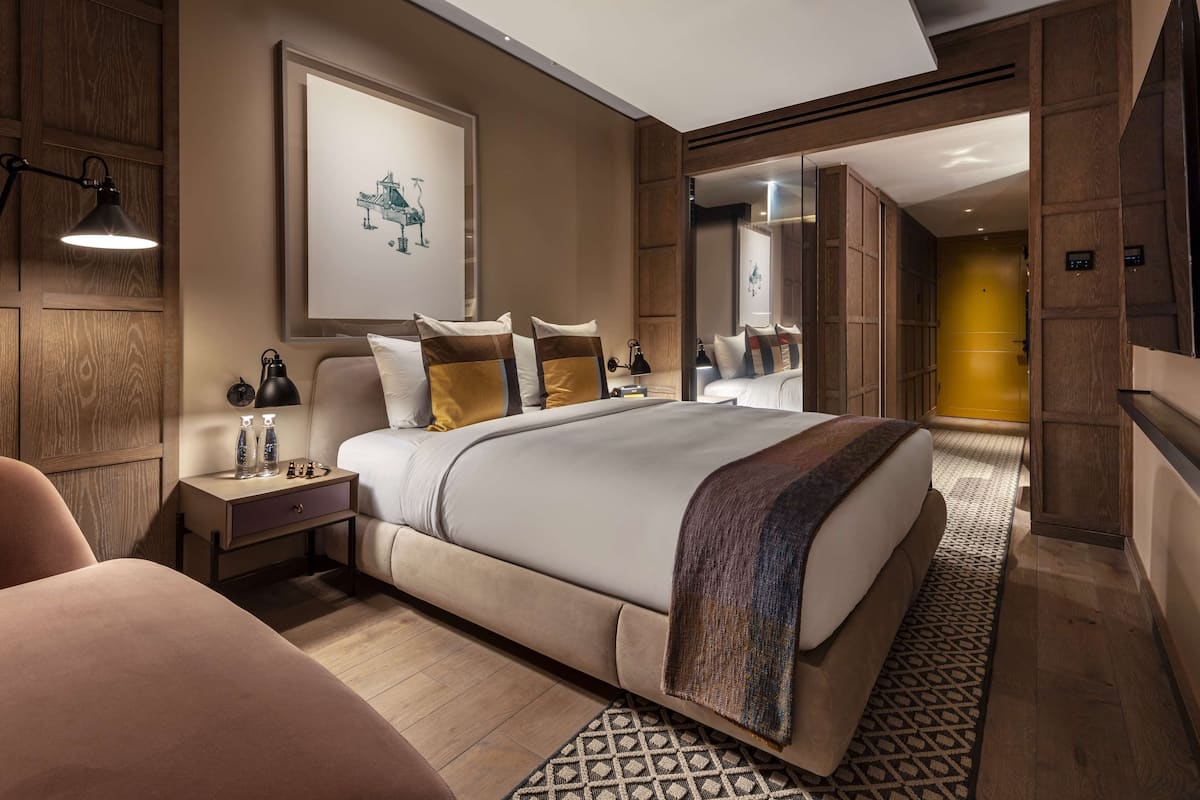 A modern hotel room with a large bed, a nightstand with a lamp, a framed picture on the wall, and a view of the hallway.