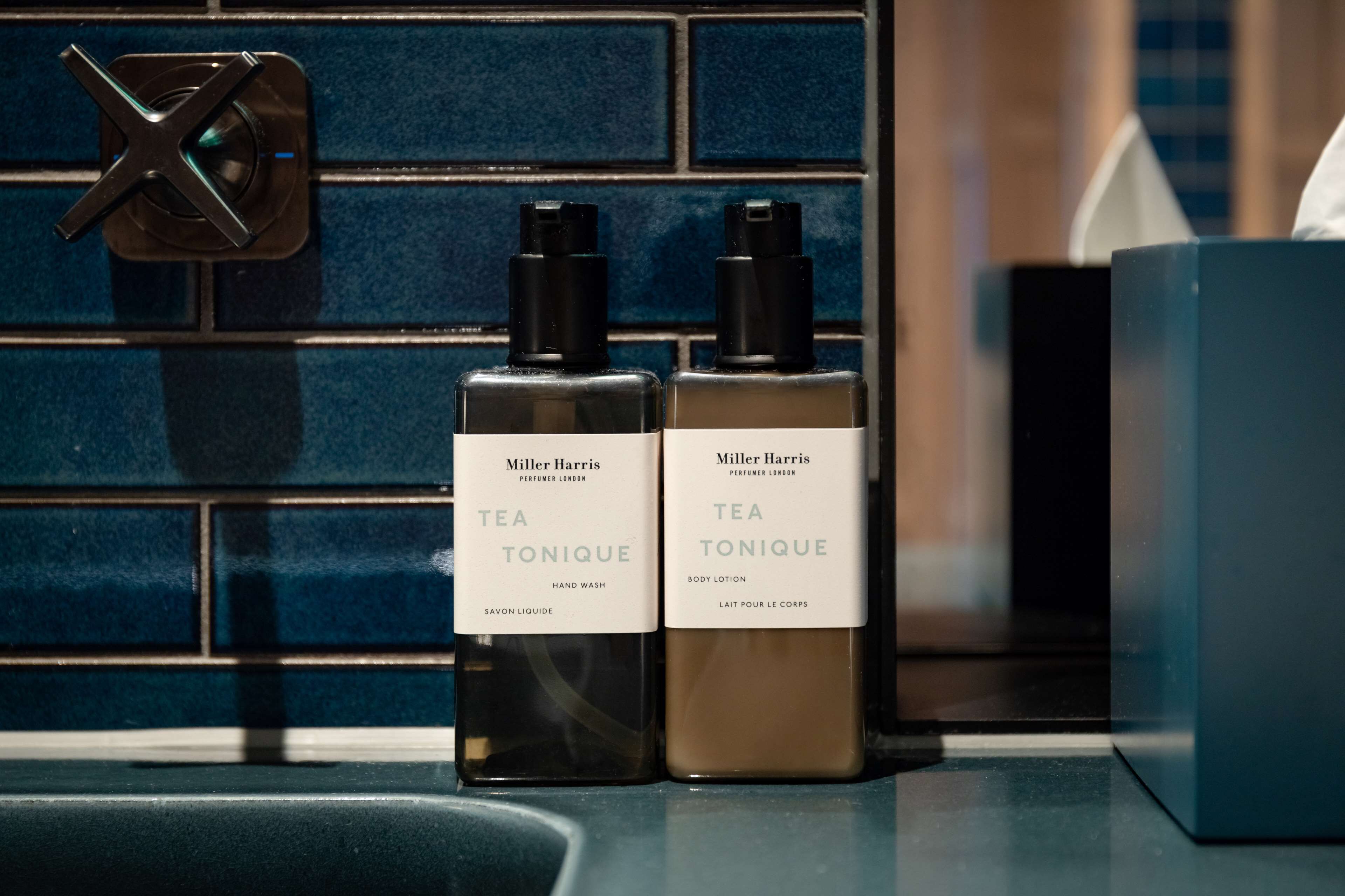 Two bottles of Miller Harris Tea Tonique hand wash and body lotion on a bathroom counter with a blue tiled backsplash.