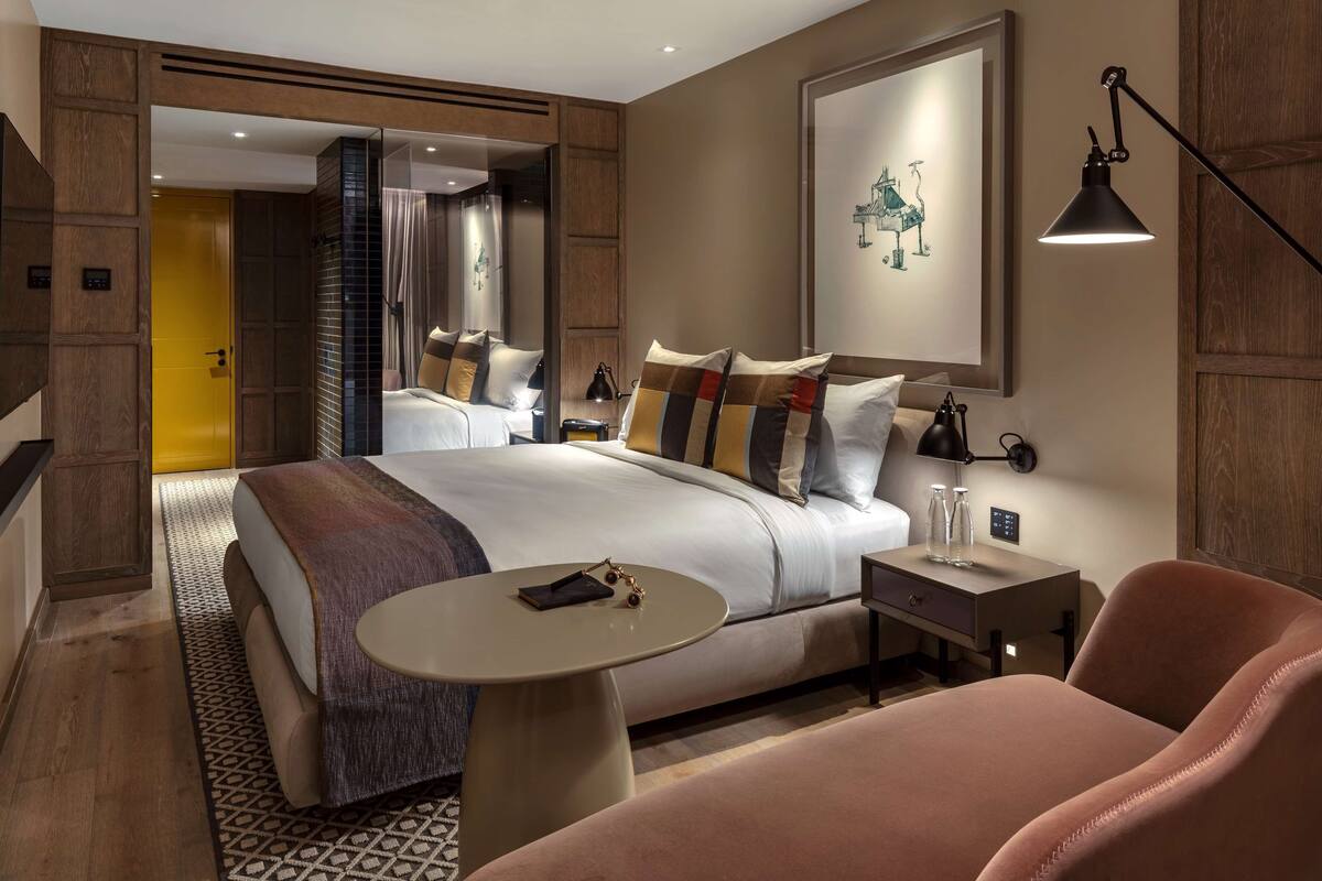 A modern hotel room with a large bed, a small round table, a chair, and a desk with a lamp.