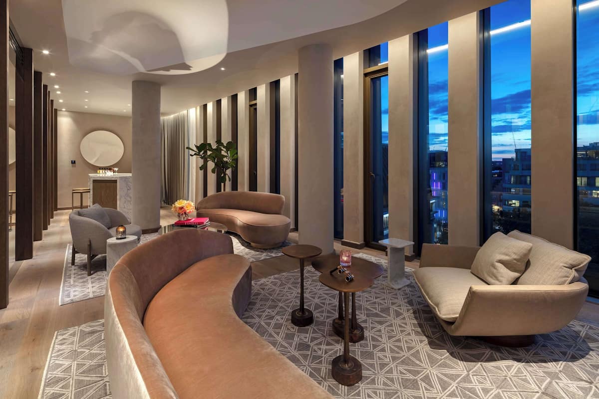 A modern hotel lobby with a curved sofa, armchairs, a small table with a plant, and large windows offering a city view.