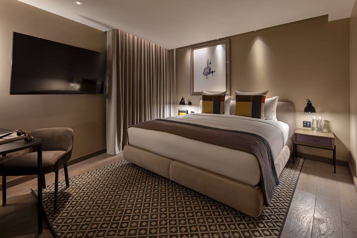 A modern hotel room with a large bed, a desk, a chair, a TV, and a bedside table with a lamp.