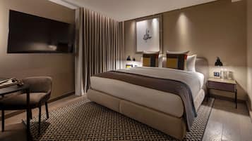 A modern hotel room with a large bed, a desk, a chair, a TV, and a bedside table with a lamp.