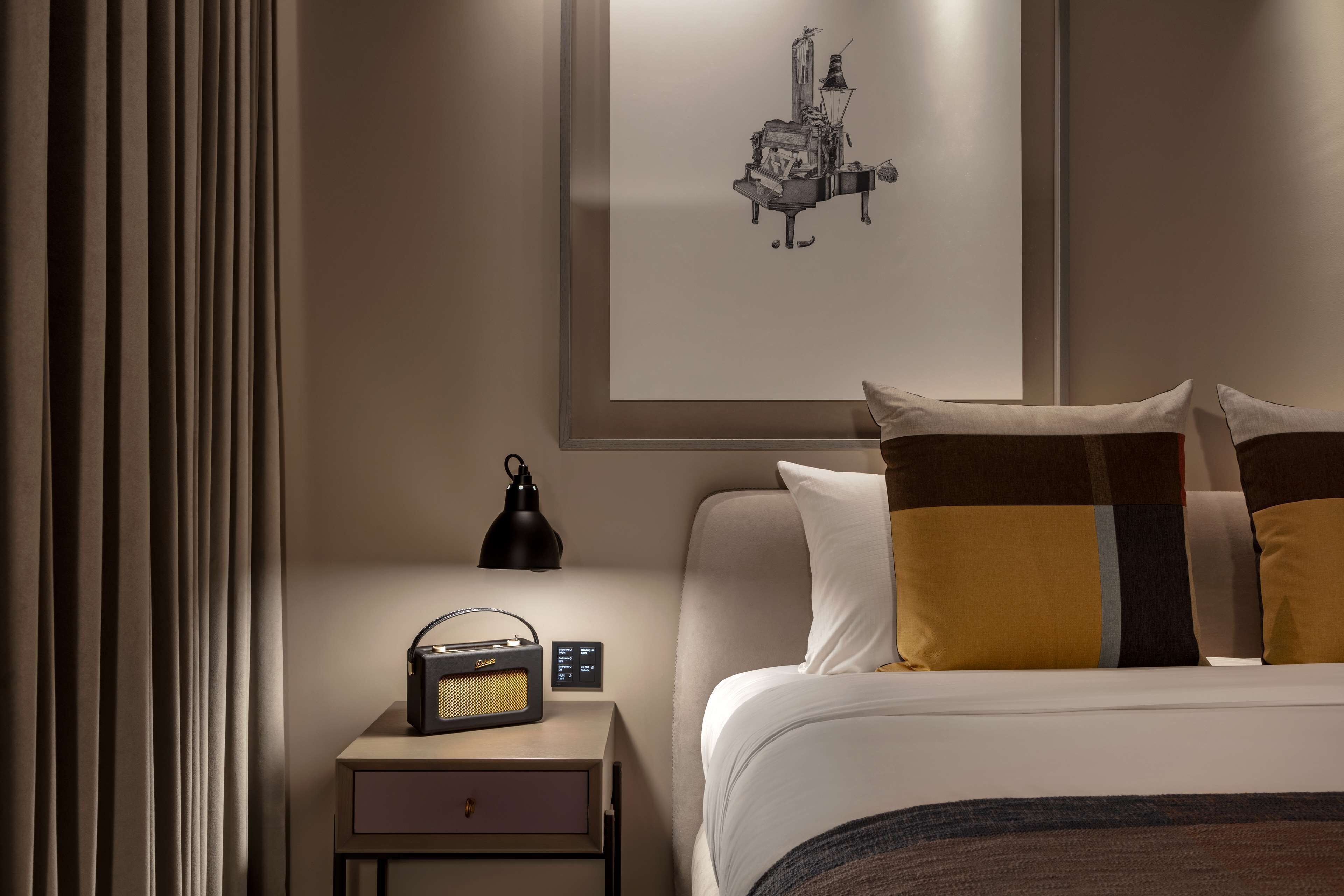 Quarto de hotel com cama, mesa de cabeceira com abajur, rádio e um quadro na parede.