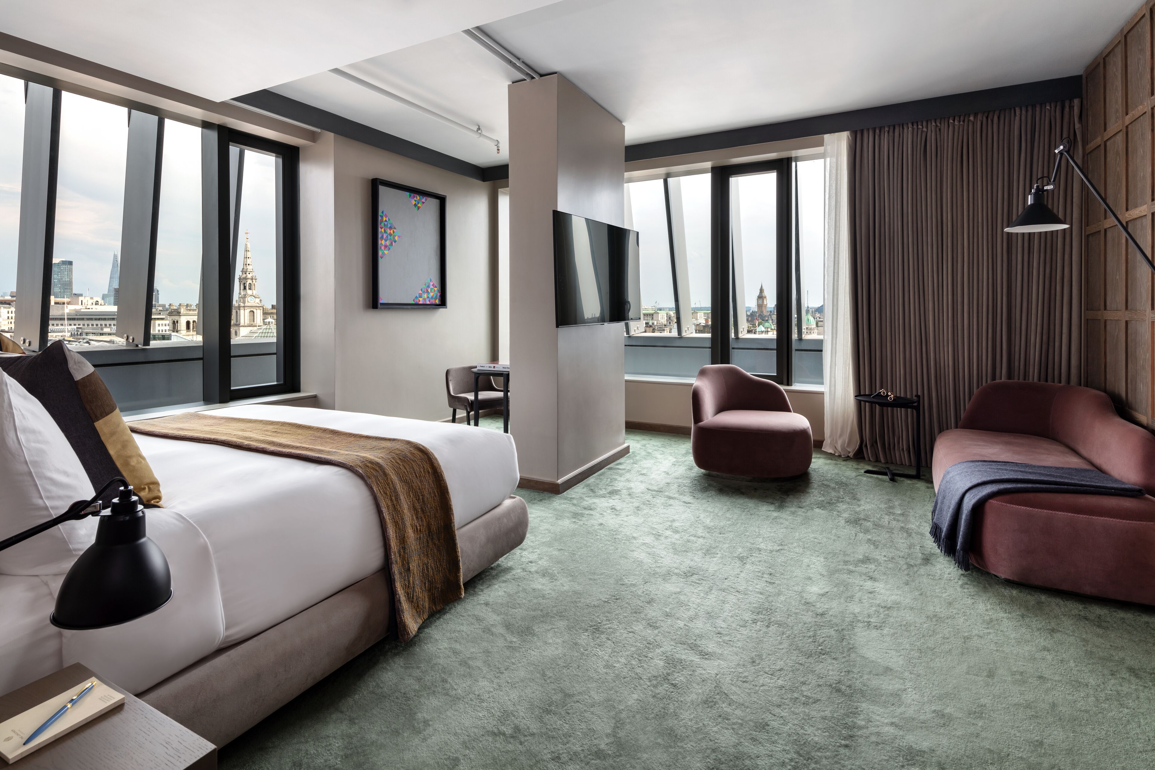 A hotel room with a large bed, a desk, a chair, a sofa, a TV, and large windows with a city view.