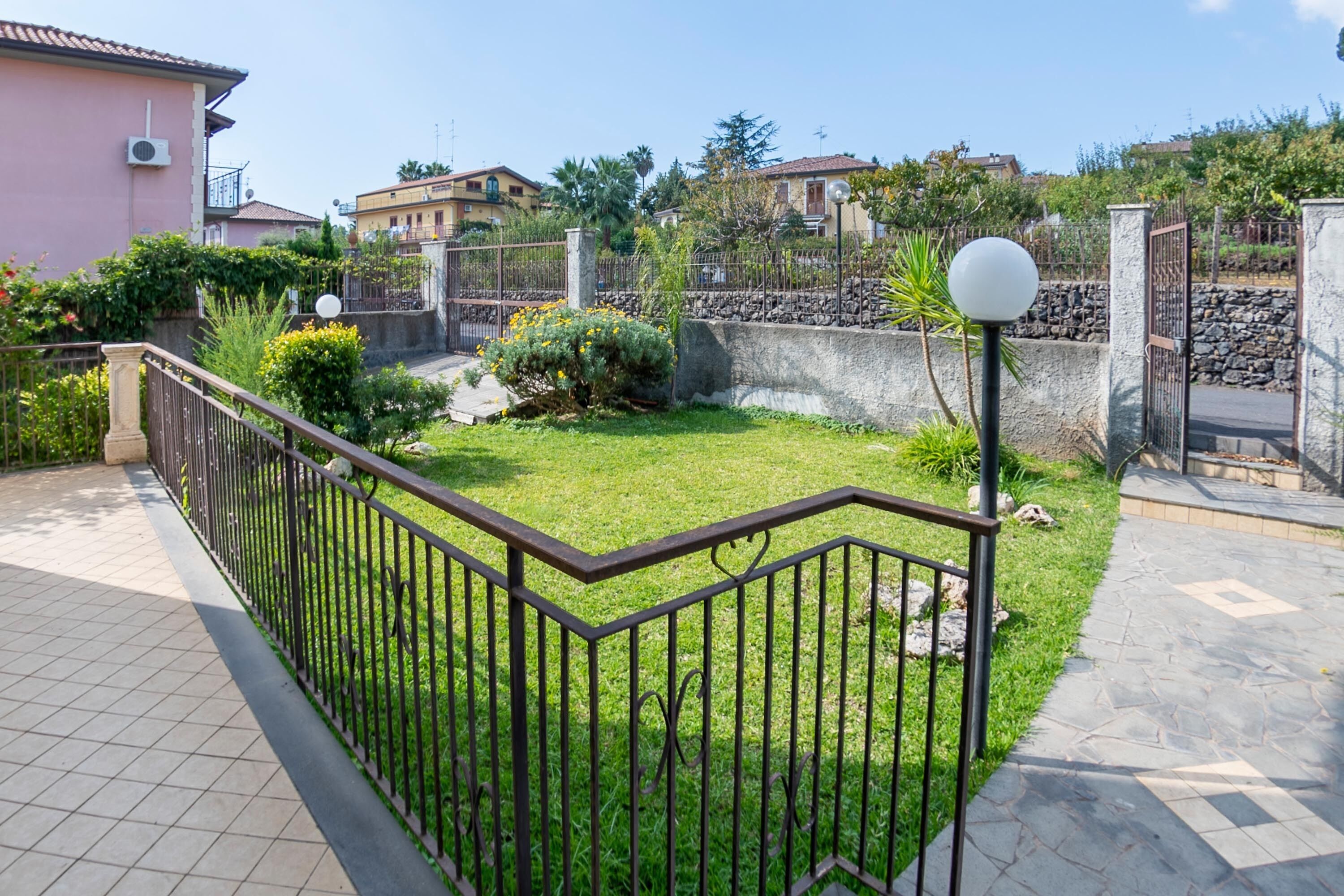 A residential area with a paved walkway, a metal fence, and a grassy lawn.