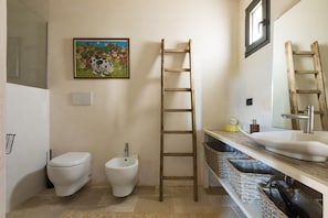 A bathroom with a toilet, bidet, wooden ladder, sink, and a painting on the wall. - Villa Joy (Ostuni)