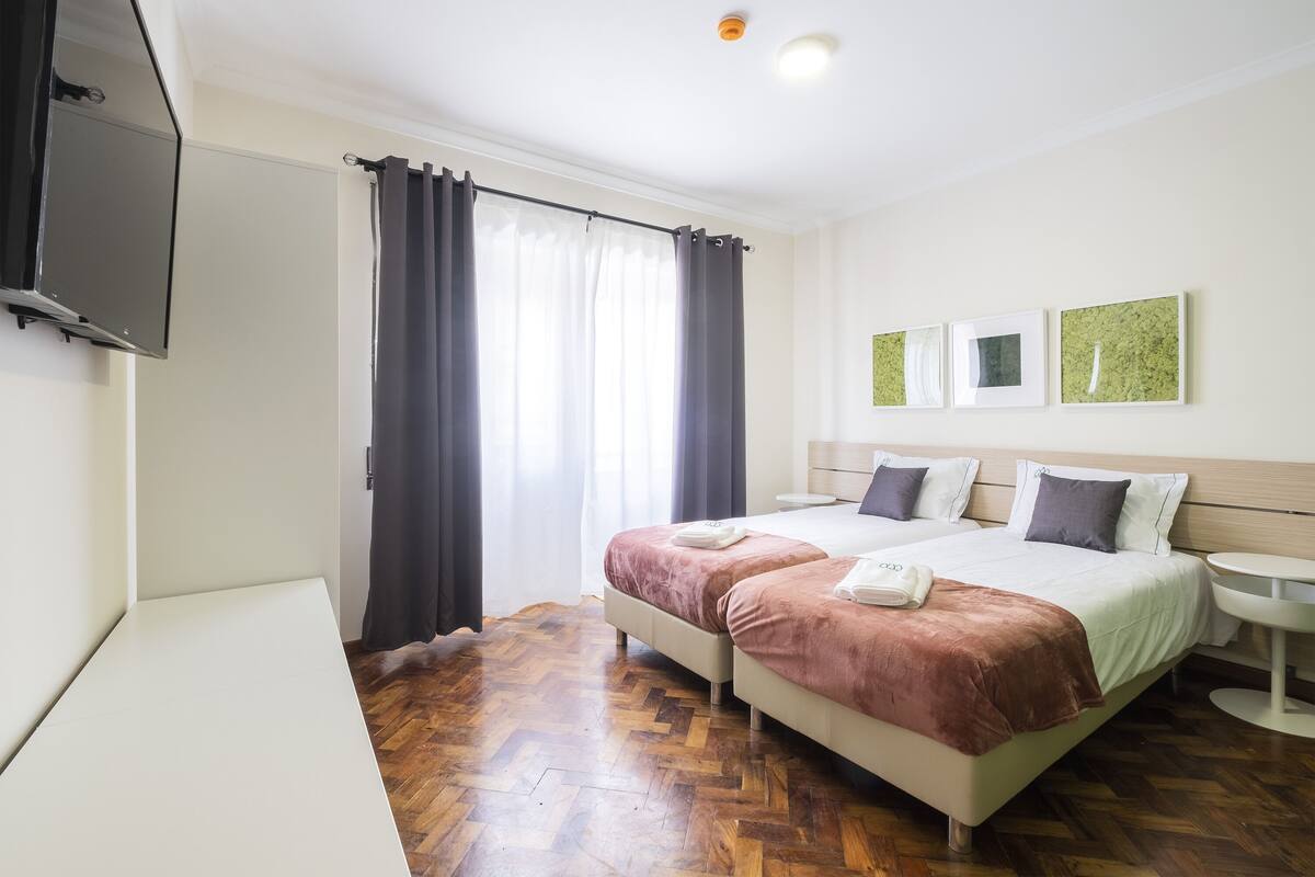 A hotel room with two beds, a flat-screen TV, a bedside table, and a window with curtains.