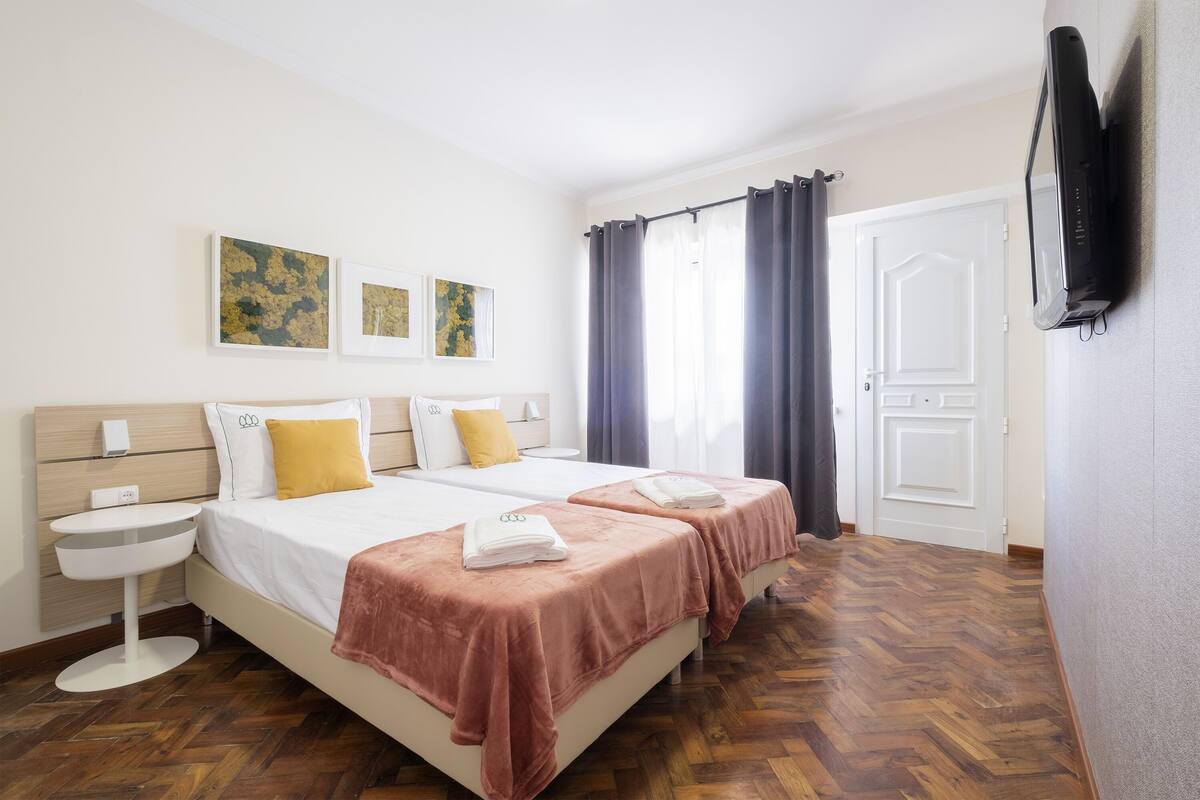 A hotel room with two beds, a bedside table, a flat-screen TV, and artwork on the wall.
