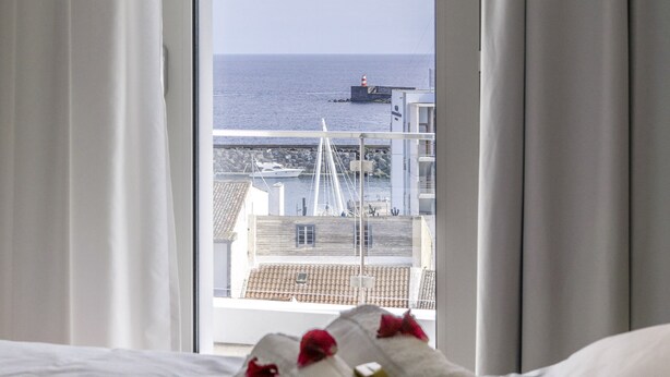 Ett hotellrum med en säng, ett fönster med gardiner och utsikt över havet och byggnaderna.