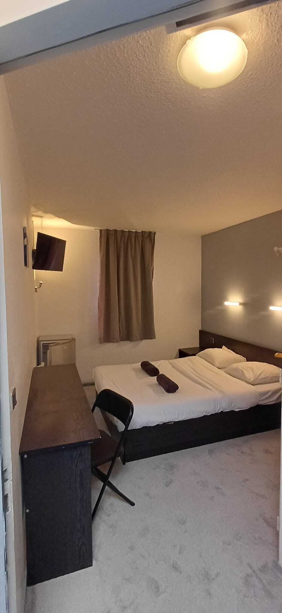 Ein kleines Hotelzimmer mit einem Einzelbett, einem Schreibtisch, einem Stuhl, einem flachbildschirmfernseher an der Wand und einer Deckenleuchte.