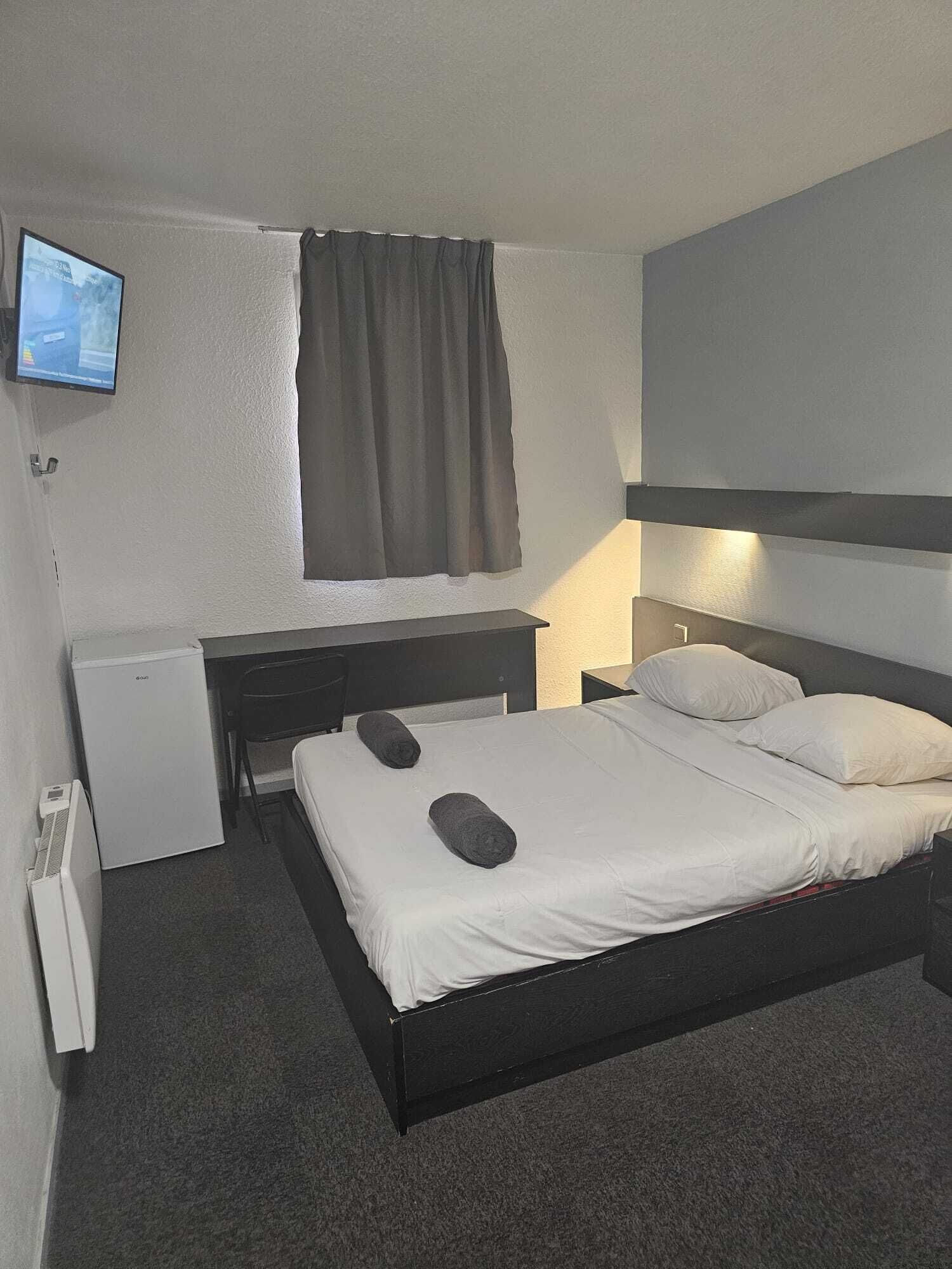 A hotel room with a bed, a desk, a chair, a TV mounted on the wall, and a window with curtains.