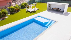 En swimmingpool med en hvid terrasse, et lille haveområde med en hvid pergola og liggestole, samt et overdækket siddeområde med bord og stole.