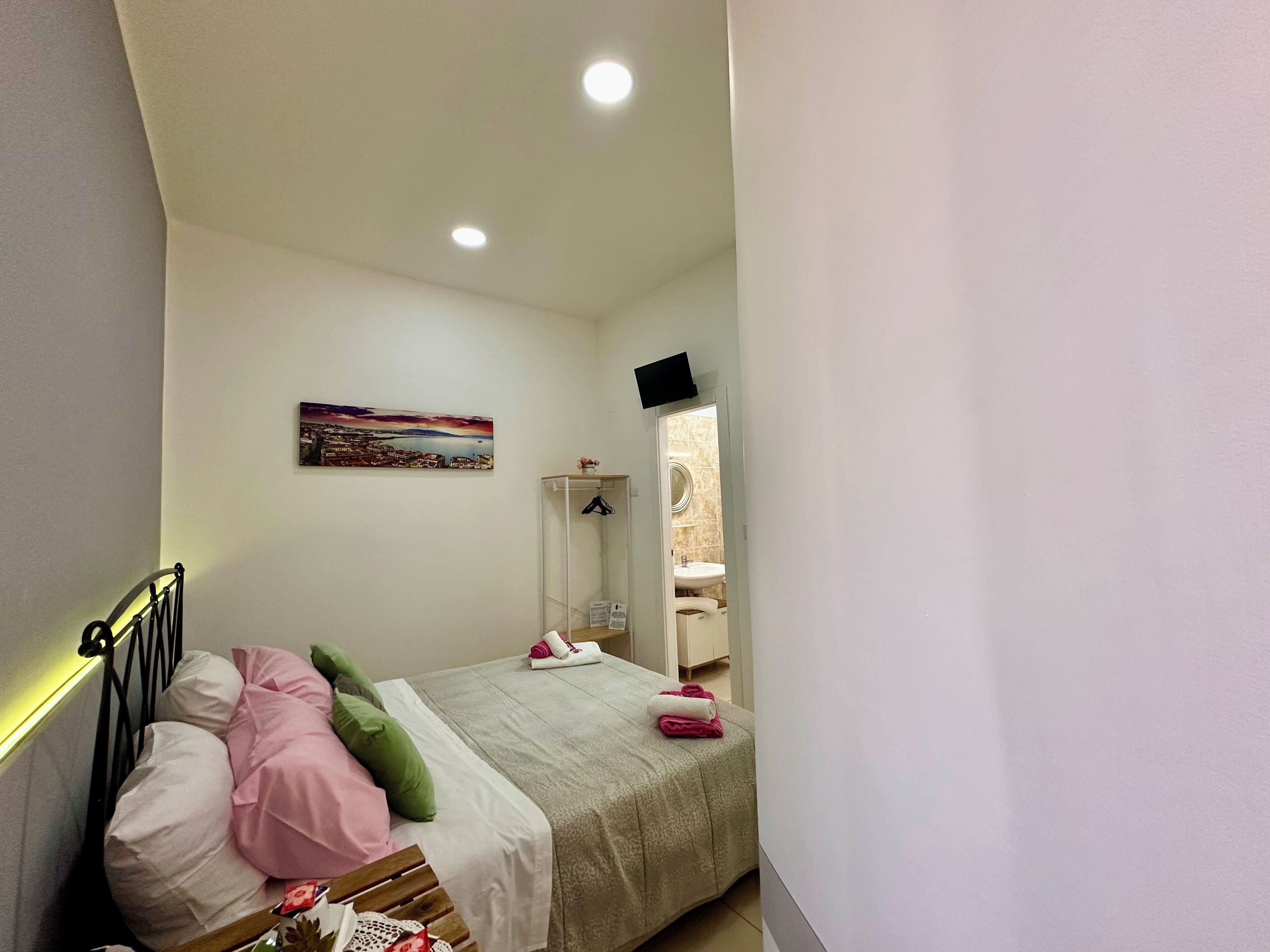 Un dormitorio con cama, almohadas, una mesita de noche, un televisor montado en la pared, un cuadro y un baño visible a través de una puerta abierta.