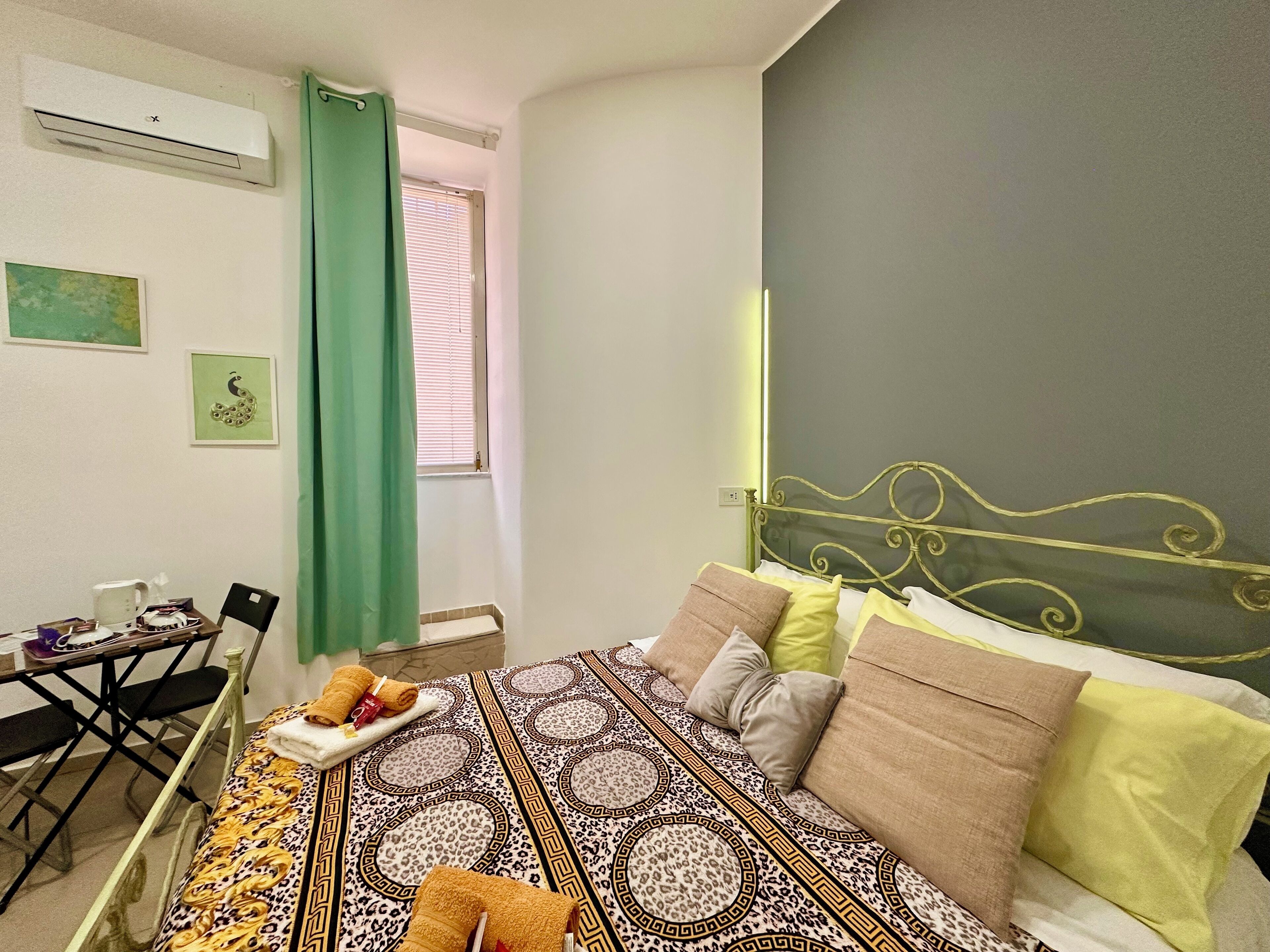 A bedroom with a patterned bedspread, a green curtain, a small table with a kettle, and a wall-mounted air conditioner.