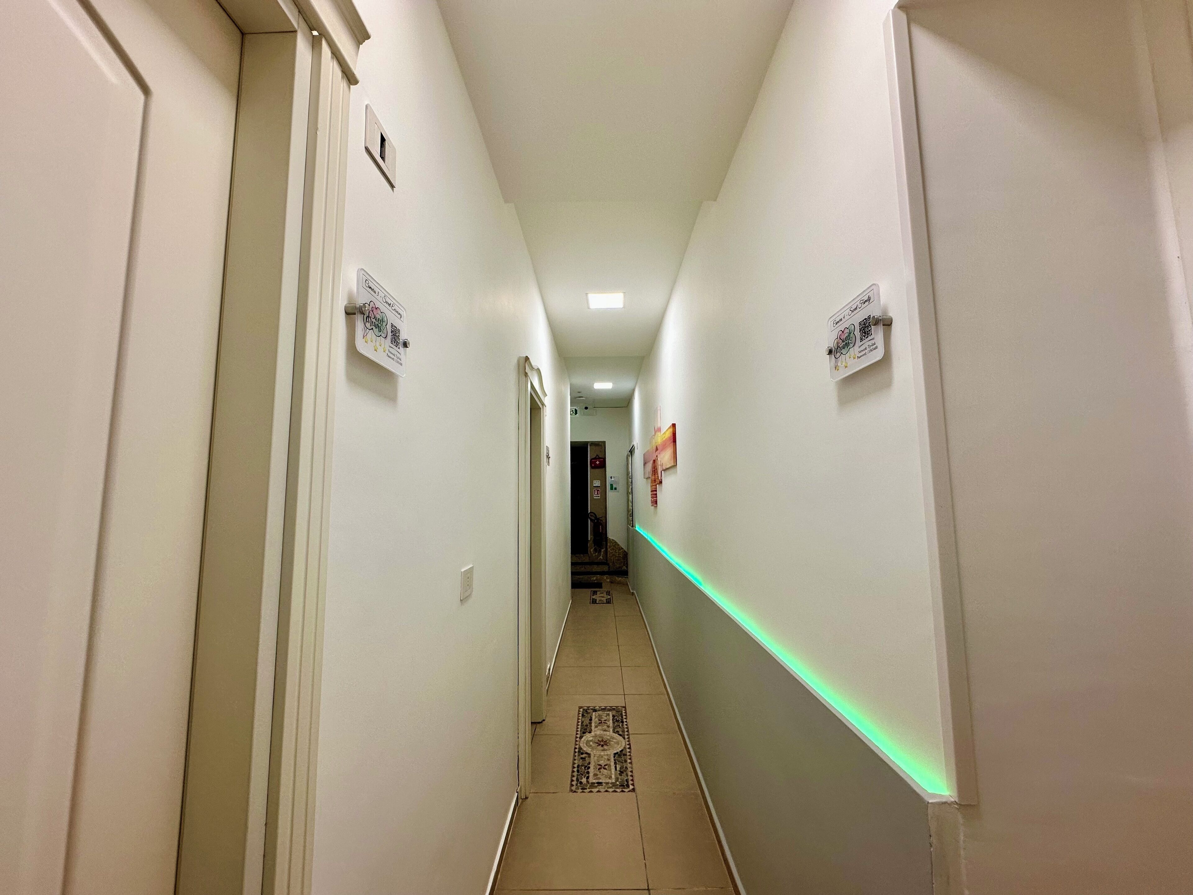 A narrow hallway with white walls, a green LED strip lighting, and a patterned rug at the end.