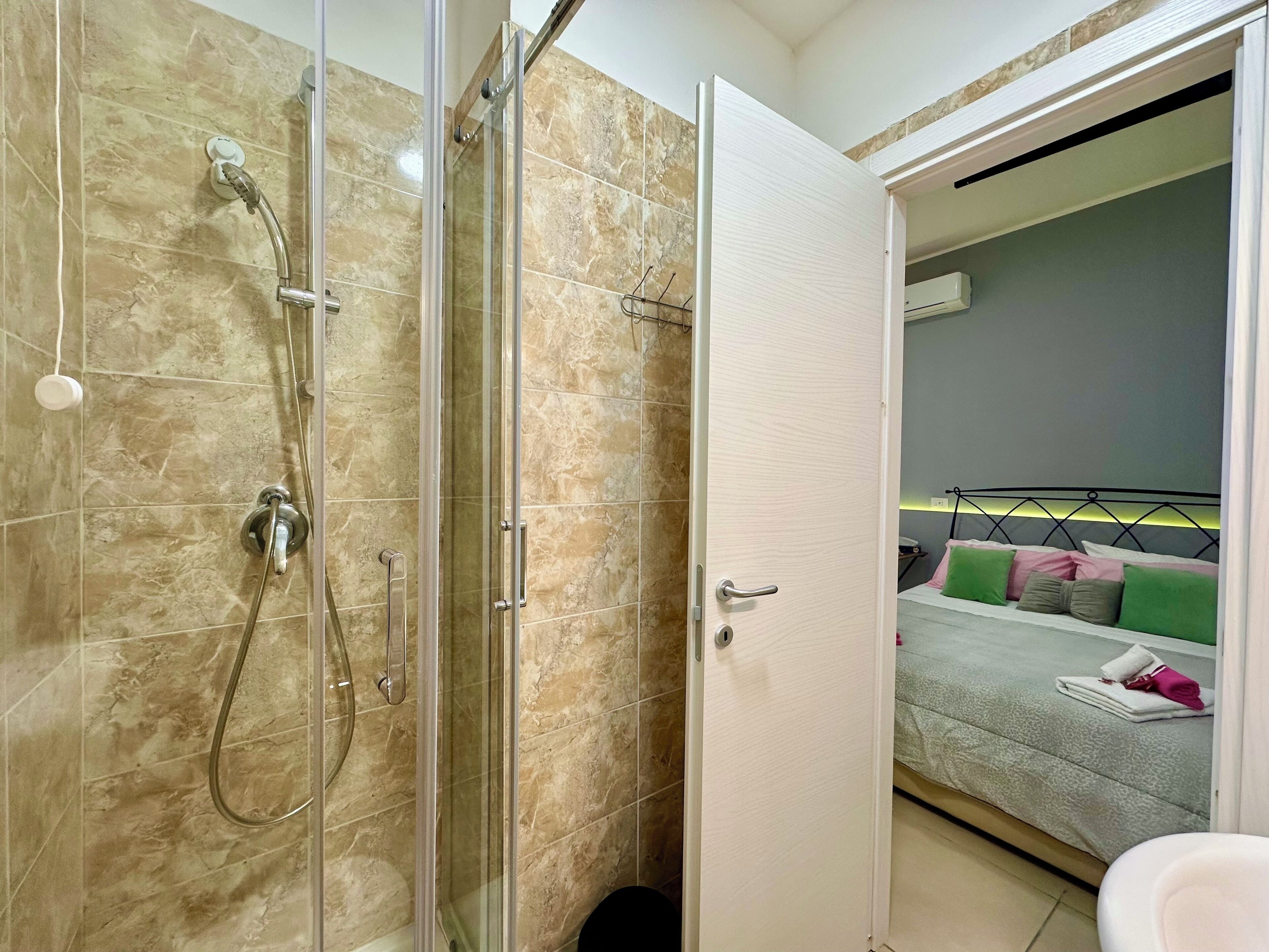 A bathroom with a glass-enclosed shower, a white door, and a bed with a patterned bedspread and pillows in the adjacent room.