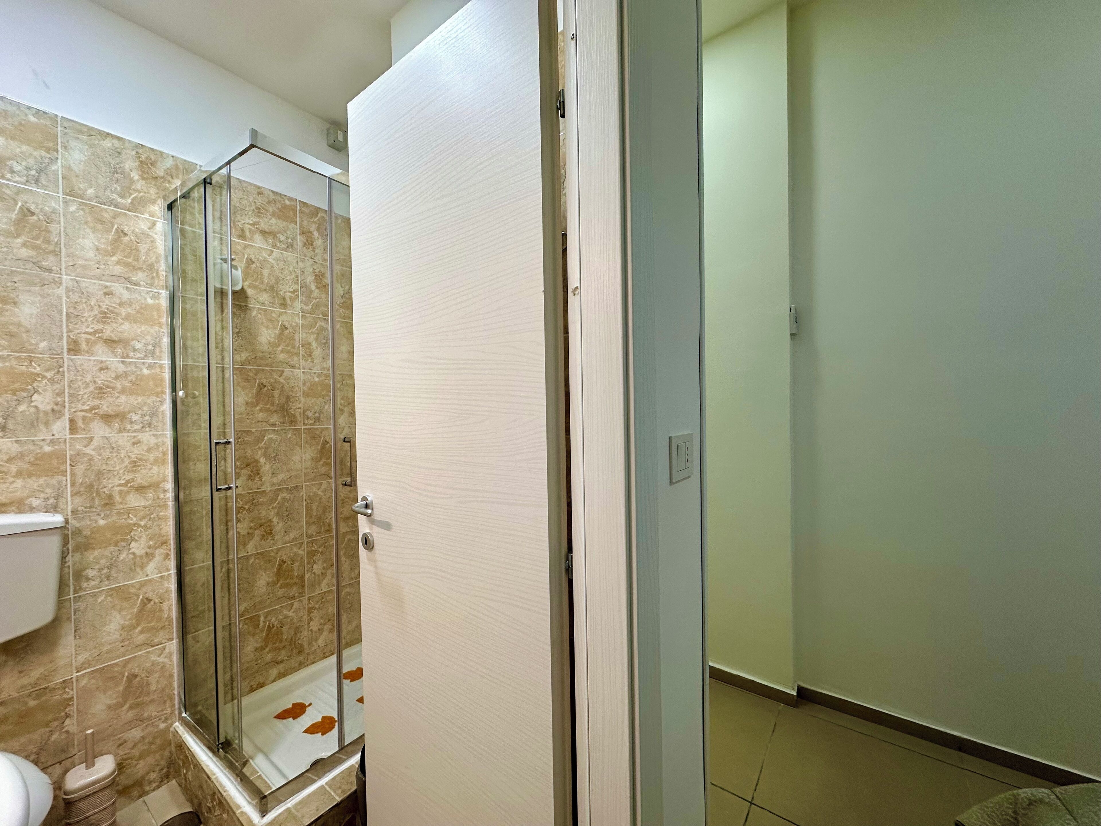 A bathroom with a glass shower enclosure, a toilet, and a door leading to another room.