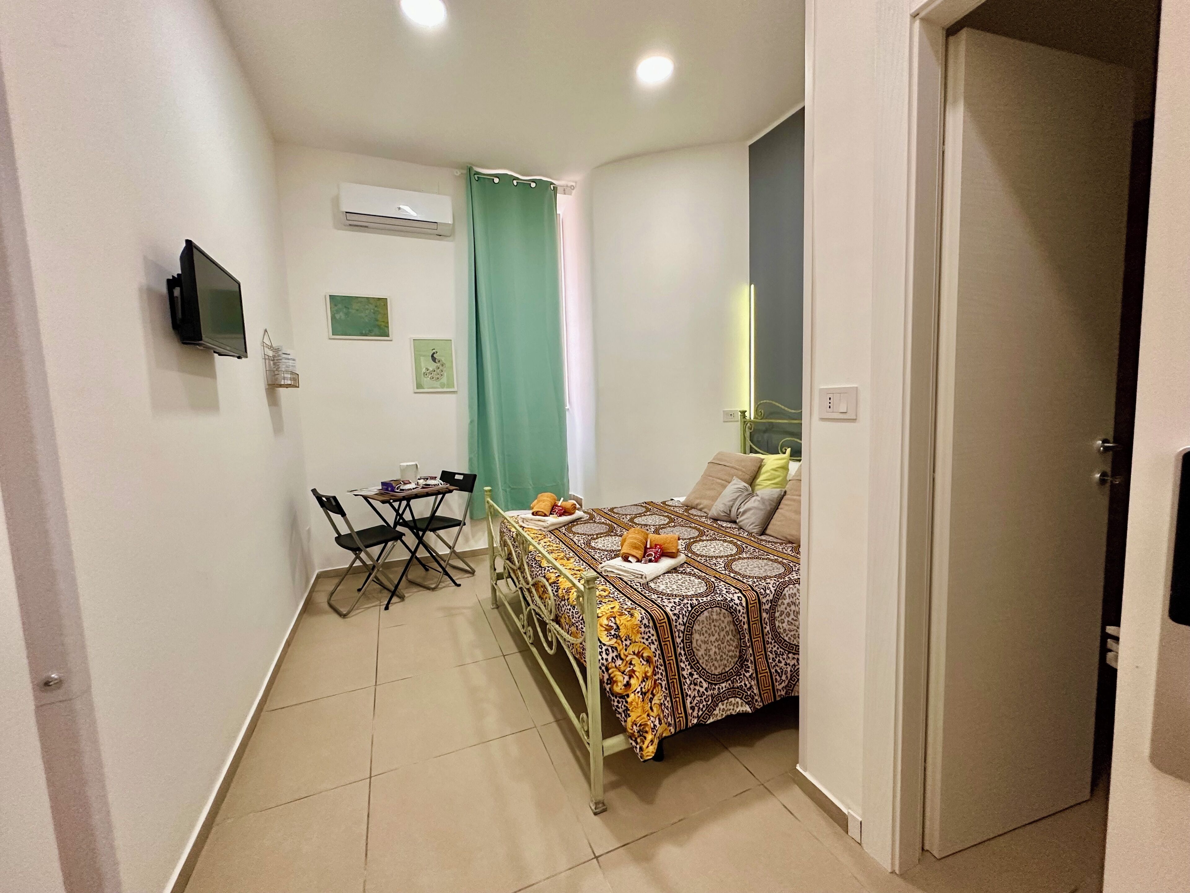 A small, well-lit room with a bed, a desk, and a chair. There is a television mounted on the wall, an air conditioning unit, and a door leading to another room.