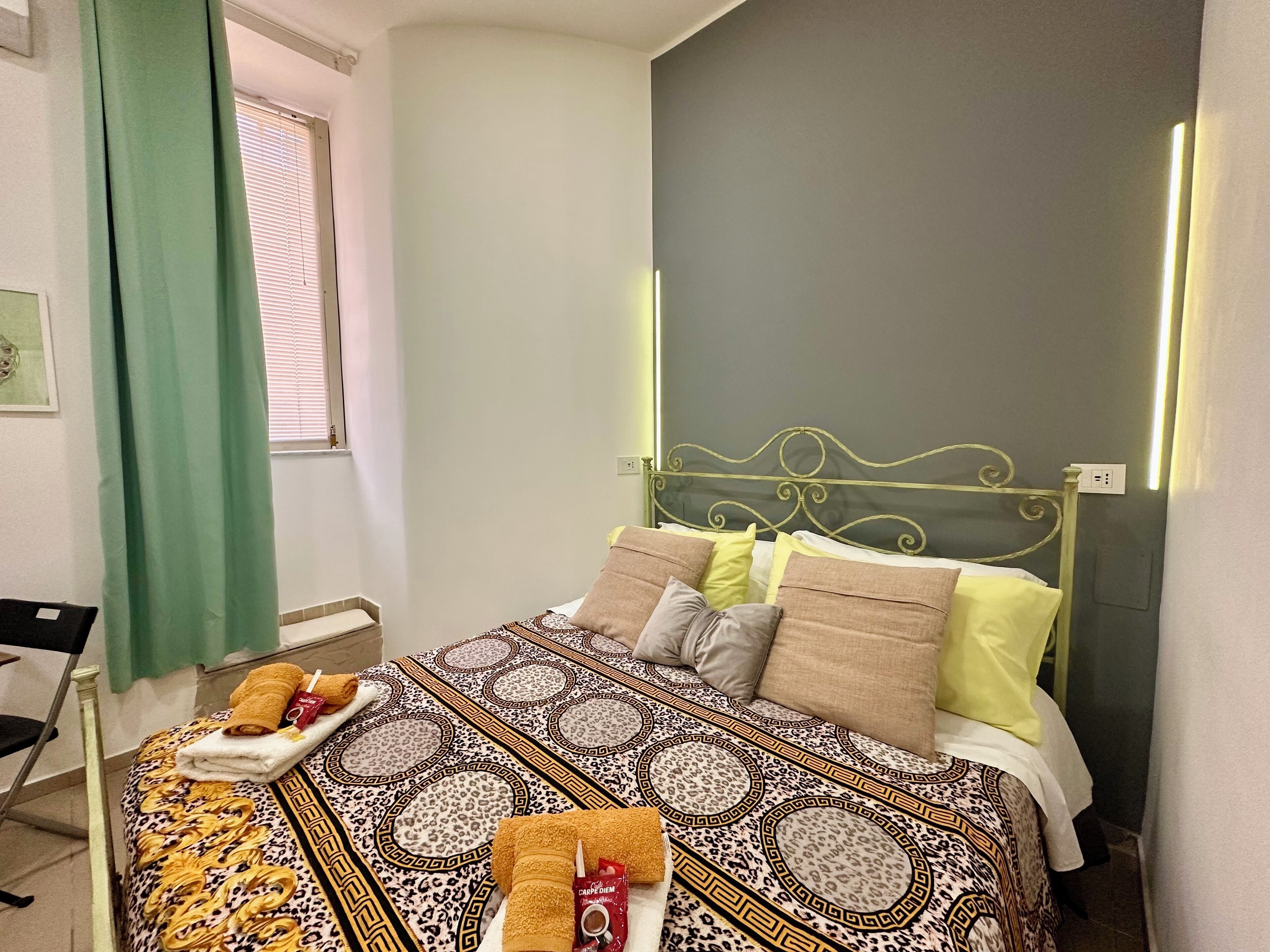 Ein ordentlich bezogenes Bett mit gemusterten Bettwäsche und mehreren Kissen, ein Nachttisch mit Handtuch und Tasse, sowie ein Fenster mit Jalousien.