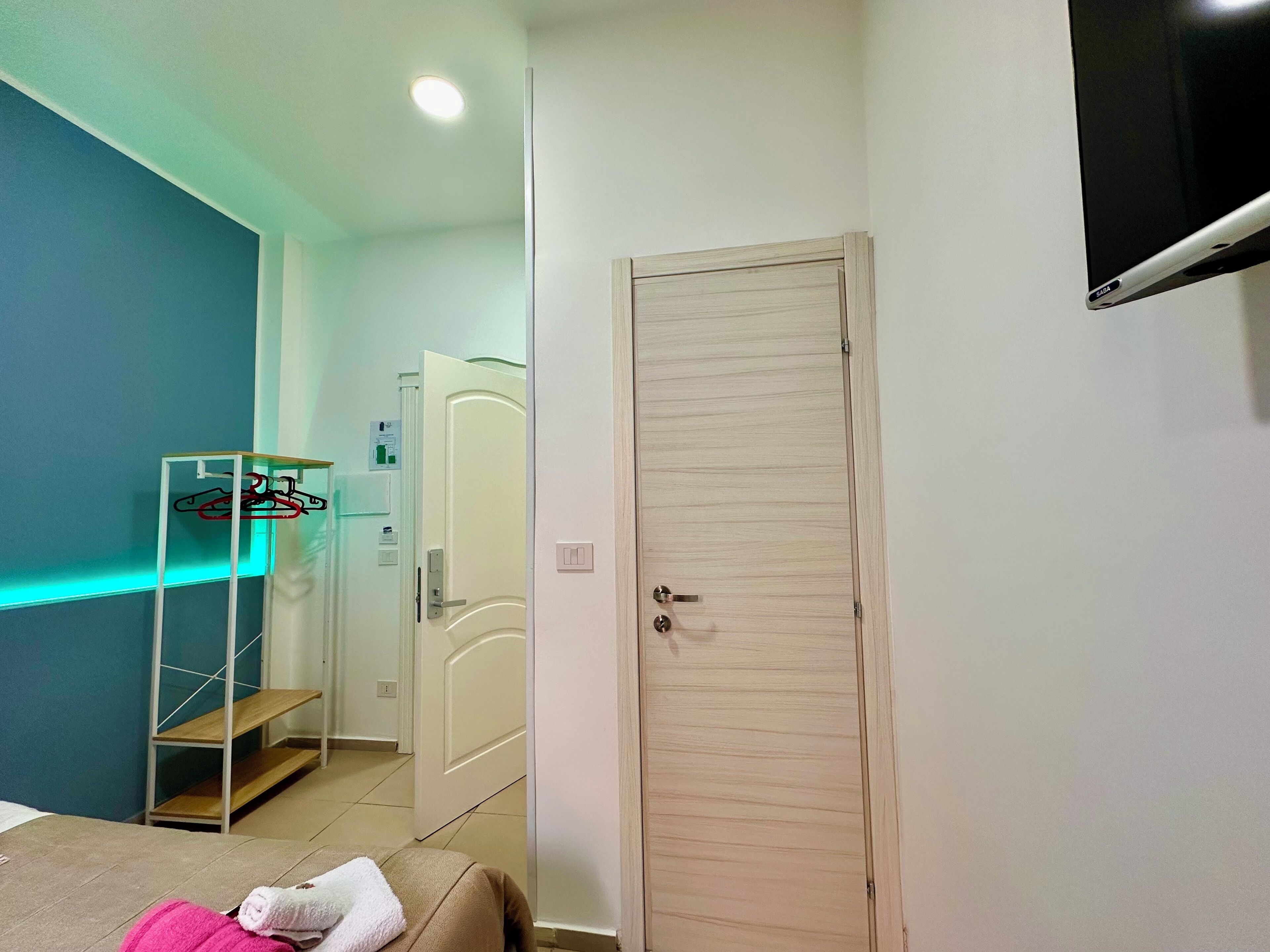 Une chambre d’hôtel avec un lit, une armoire, un miroir, une télévision et une étagère avec des vêtements.