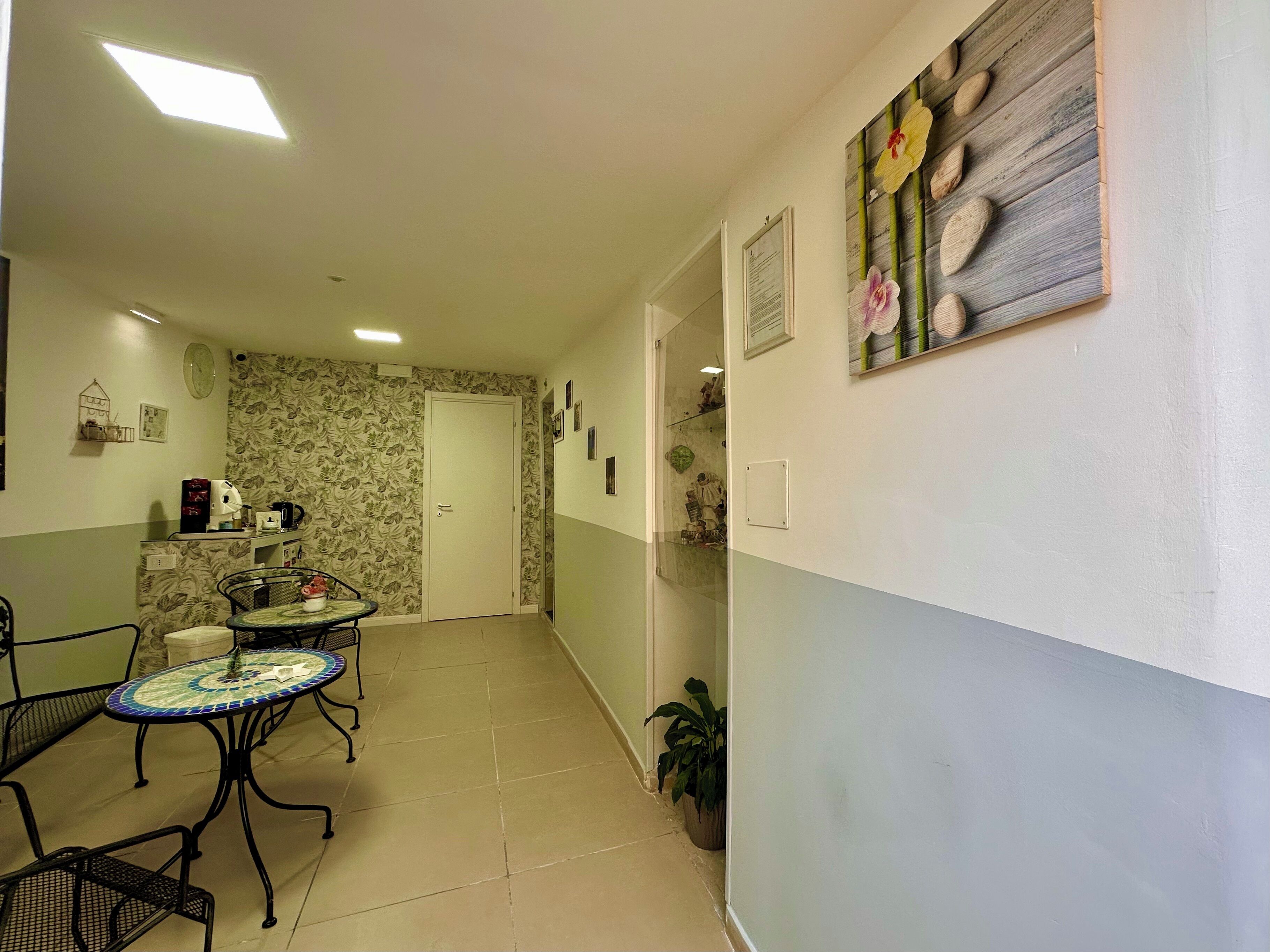 A hallway with a dining area featuring a tiled floor and metal chairs. There is a door at the end of the hallway and a wall with a floral wallpaper pattern.