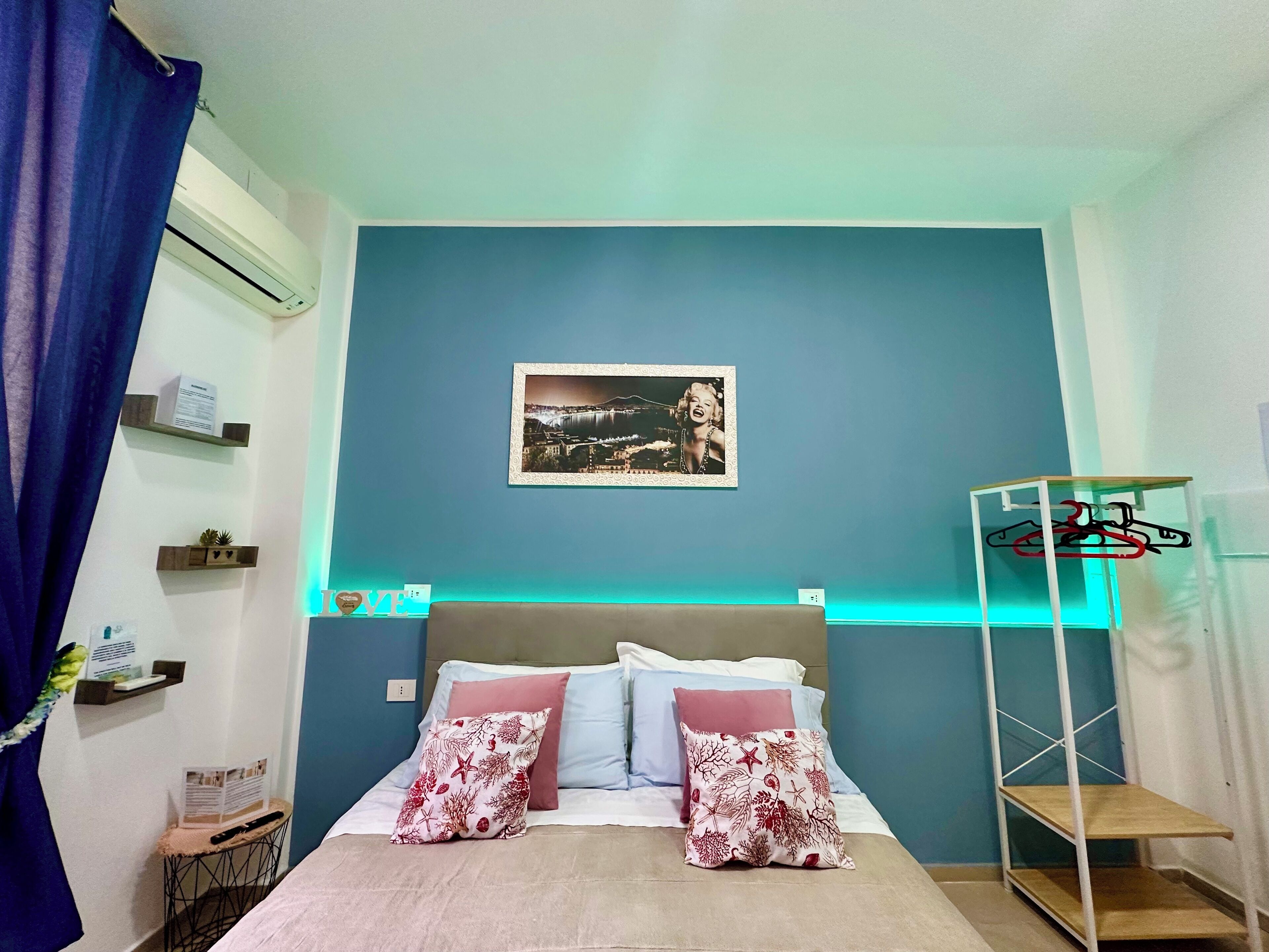Een slaapkamer met een bed, nachtkastje, een wandairconditioner, een lijst met een afbeelding en een kledingrek.
