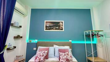 Een slaapkamer met een bed, nachtkastje, een wandairconditioner, een lijst met een afbeelding en een kledingrek.