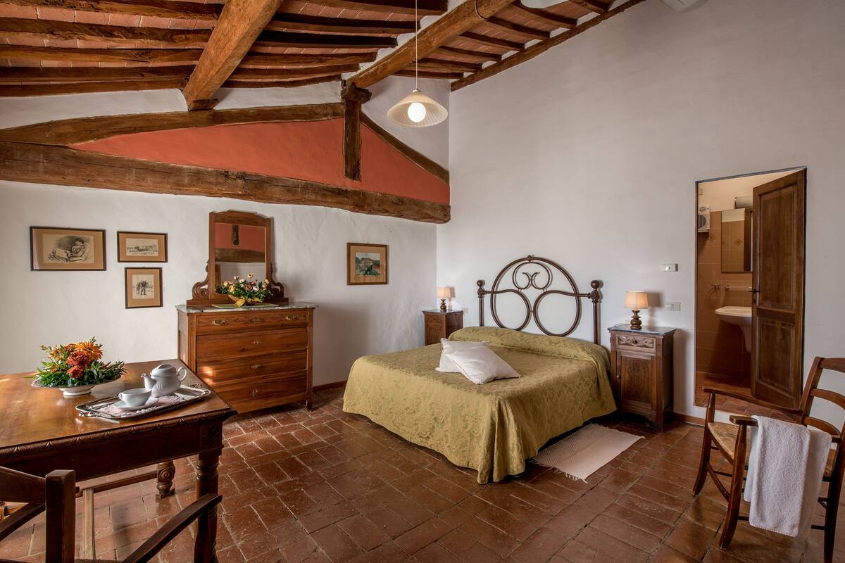 A bedroom with a wooden ceiling, a bed with a green bedspread, a dresser, a mirror, and a table with a tray of flowers.