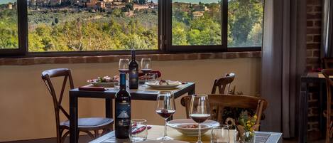 Une table à manger dressée avec des assiettes, des verres et une bouteille de vin, offrant une vue pittoresque sur un village à travers de grandes fenêtres.