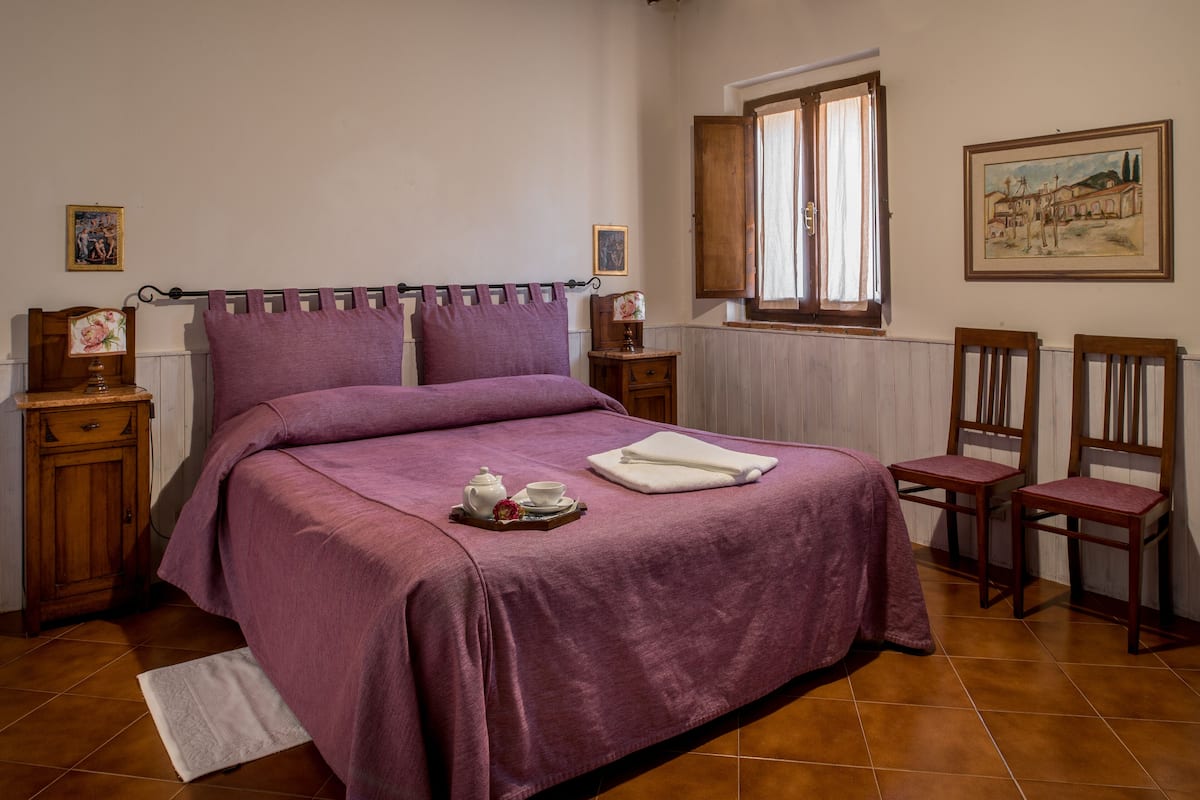A bedroom with a large bed, two bedside tables, a window with curtains, and two wooden chairs.