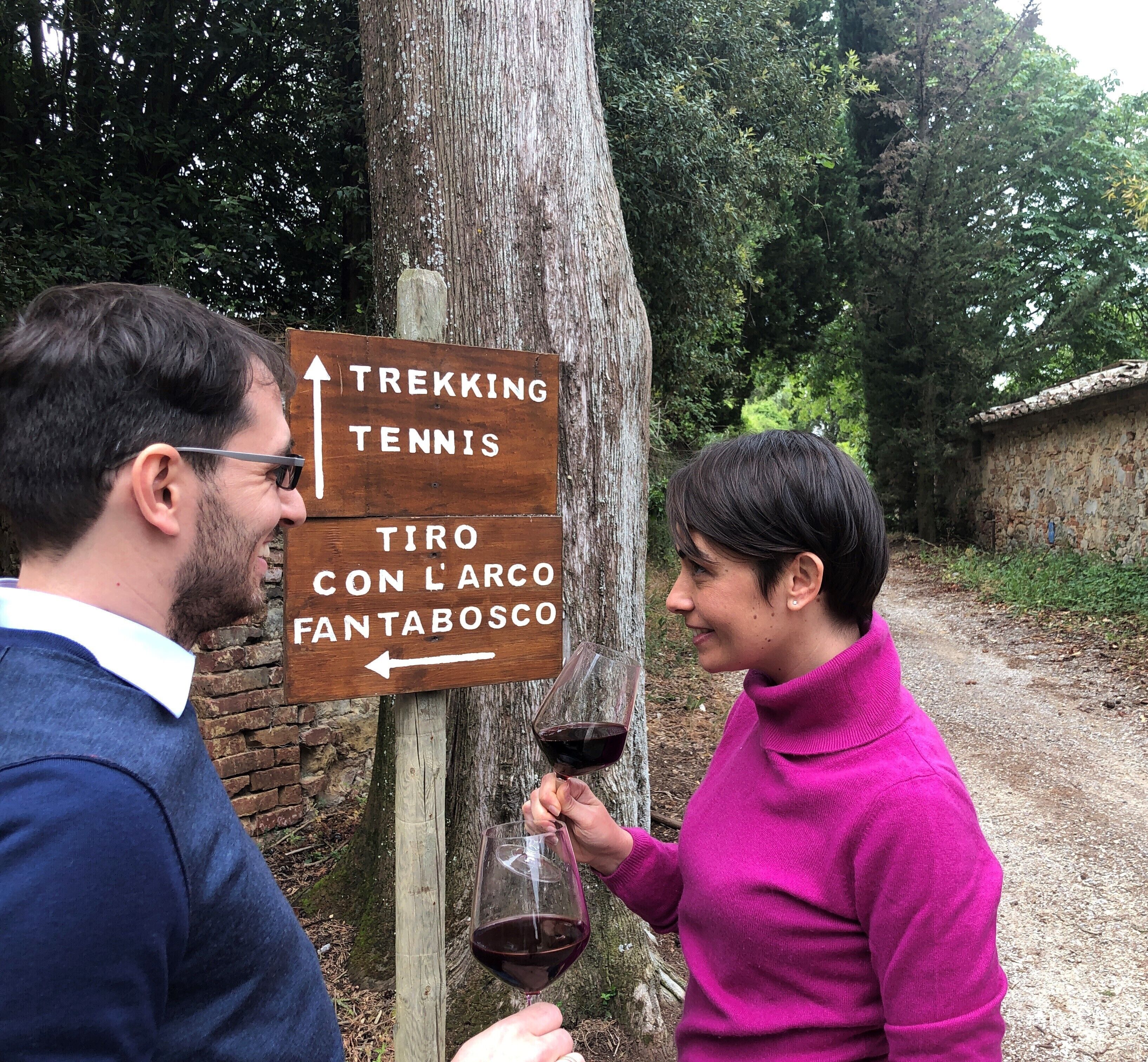 Un panneau indicateur en bois indiquant les directions pour la randonnée et le tennis, et une personne tenant un verre de vin.