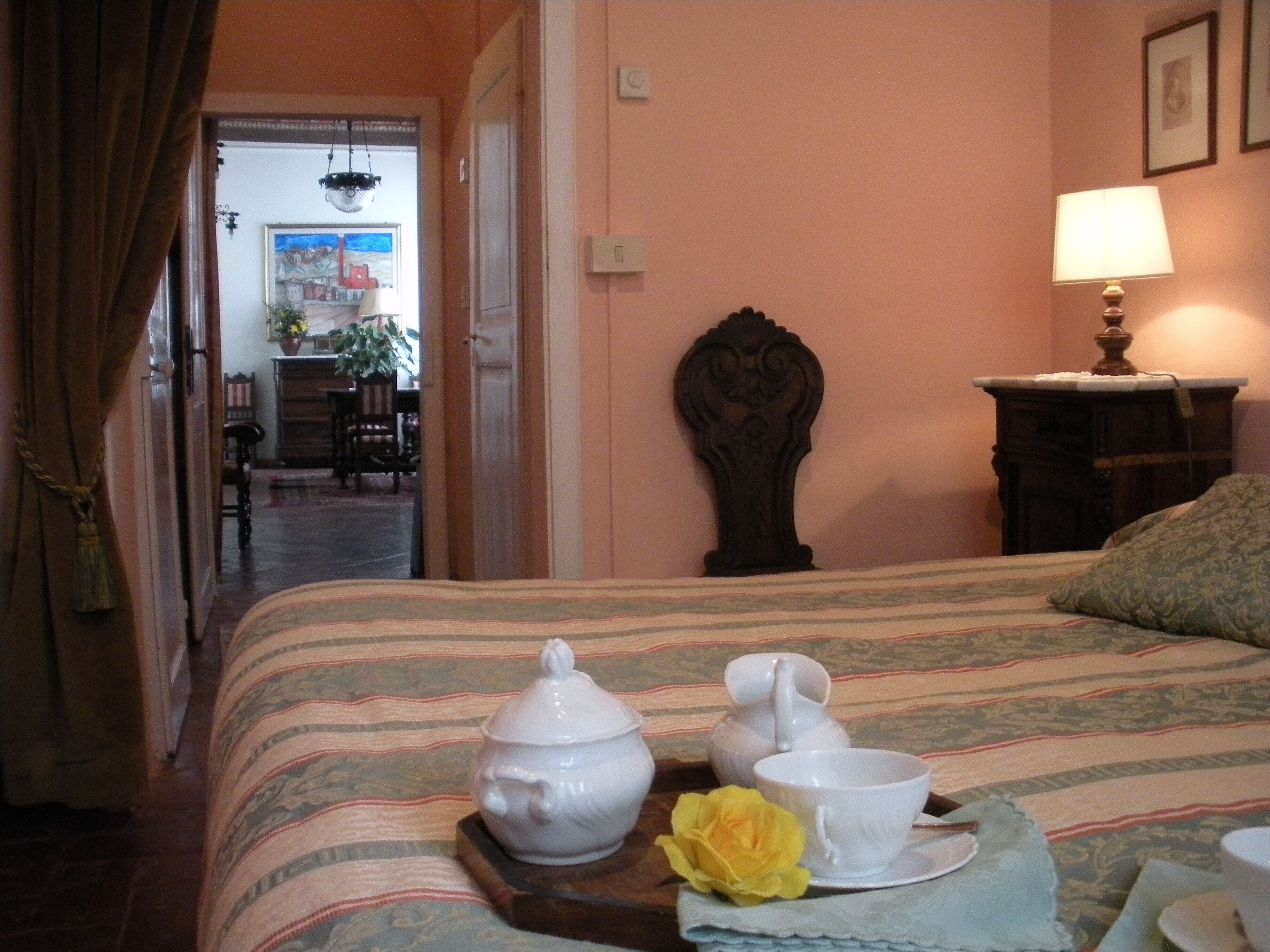 A hotel room with a bed, a bedside table with a lamp, a wooden headboard, and a view of the dining area through an open door.