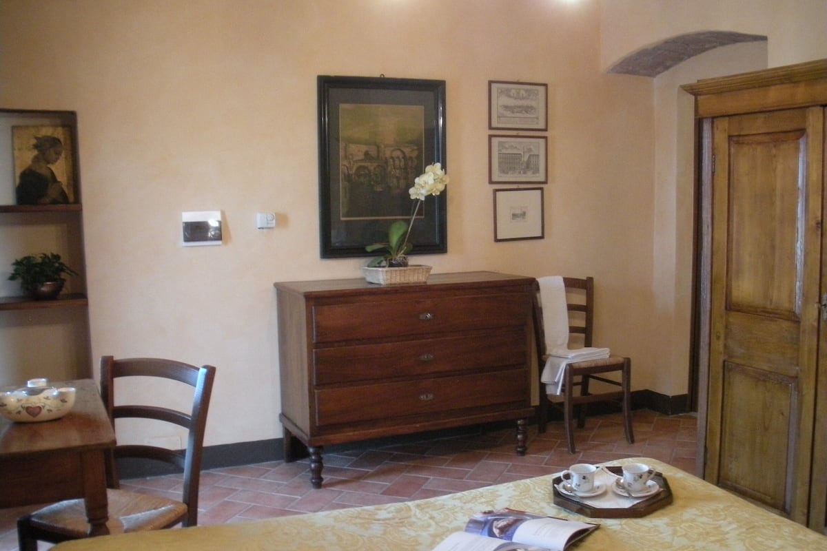 A room with a bed, a wooden dresser, a chair, a table with a tray of cups and saucers, and a framed picture on the wall.