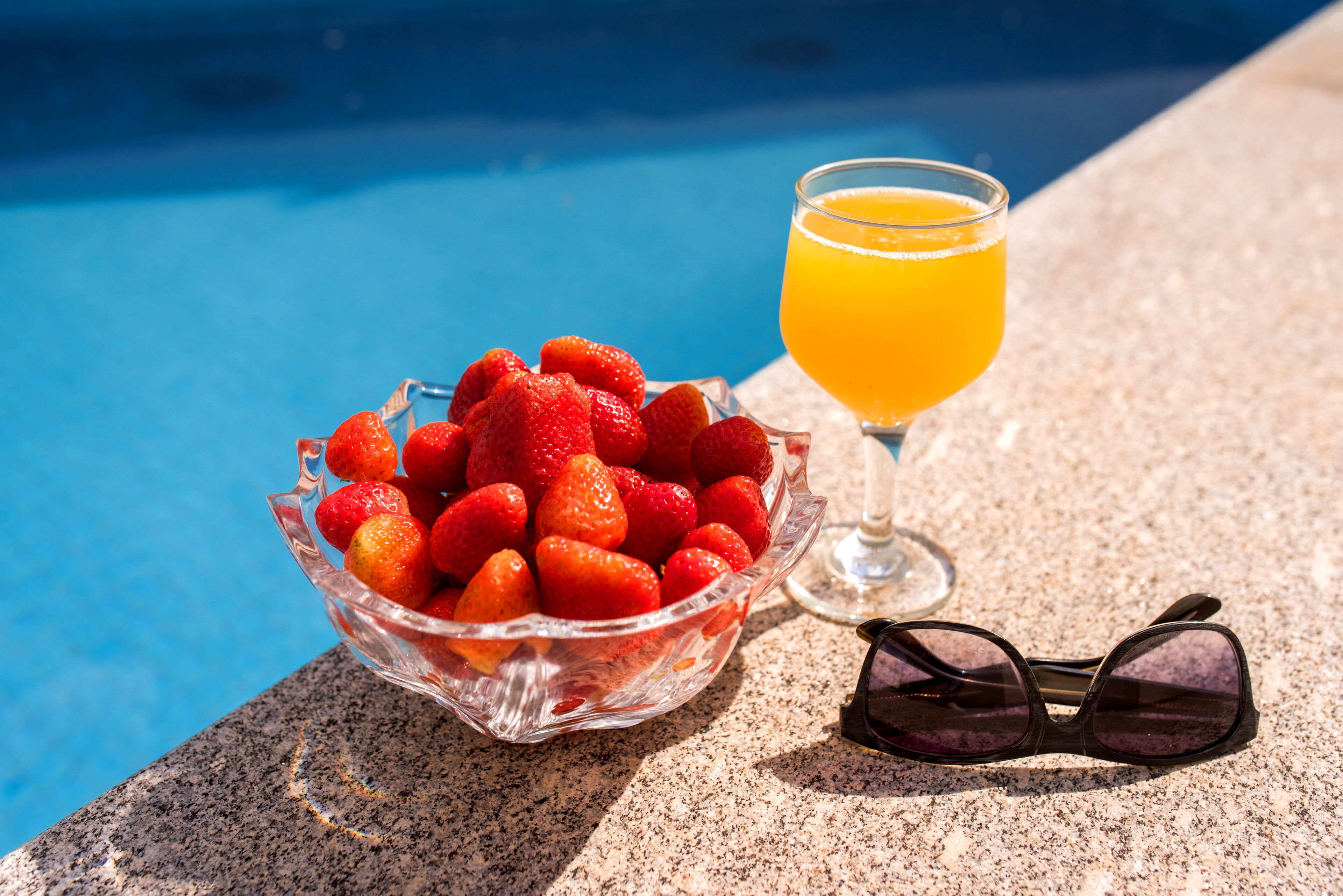 Un cuenco de vidrio con fresas, un vaso de jugo de naranja y unas gafas de sol junto a una piscina.