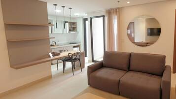 Una sala moderna con un sofá marrón, un comedor con una mesa y sillas, y una cocina al fondo.