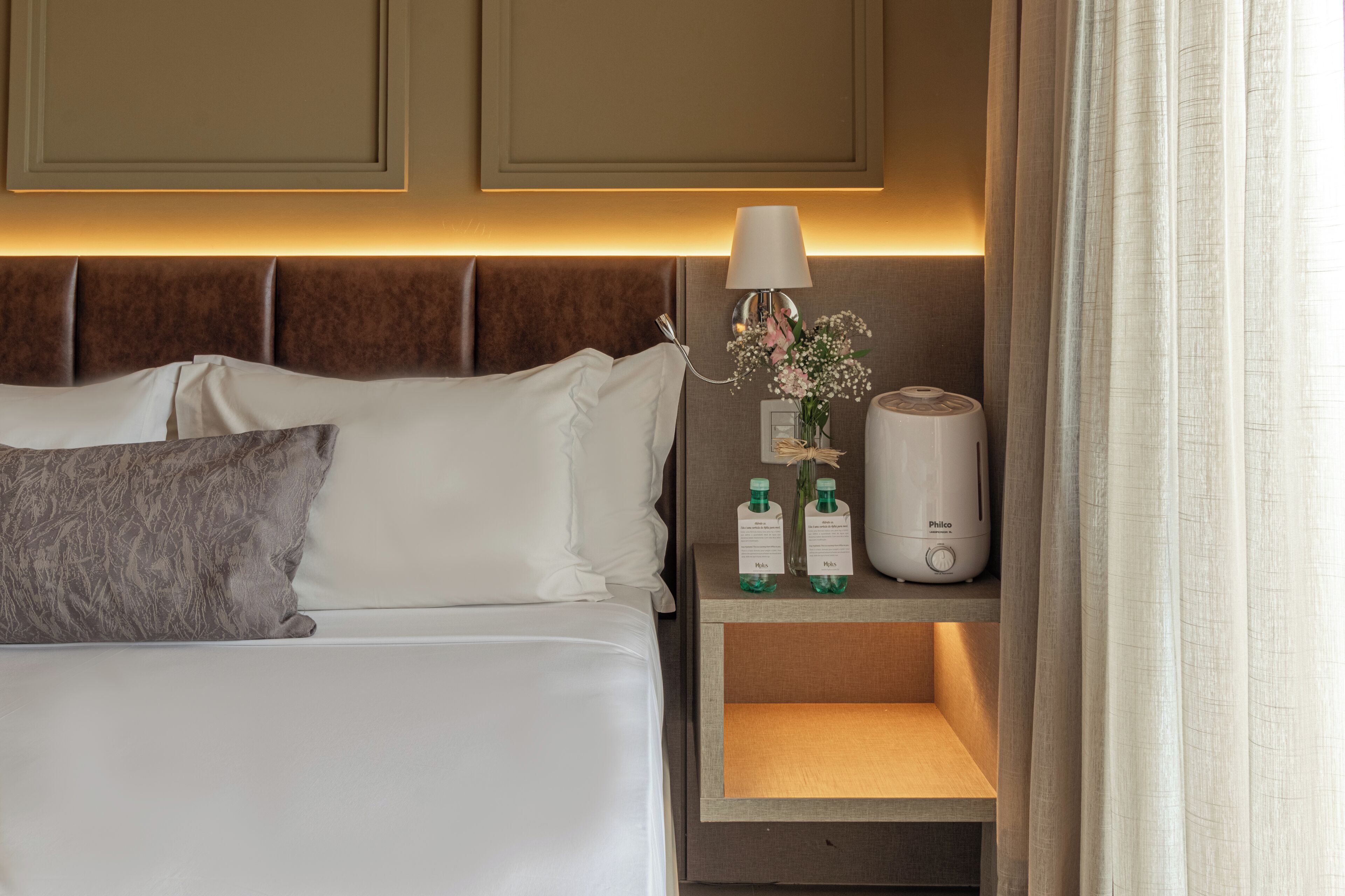 A neatly made bed with white and gray pillows, a bedside table with a lamp, two bottles of water, and a white device, possibly a humidifier, on a hotel room.