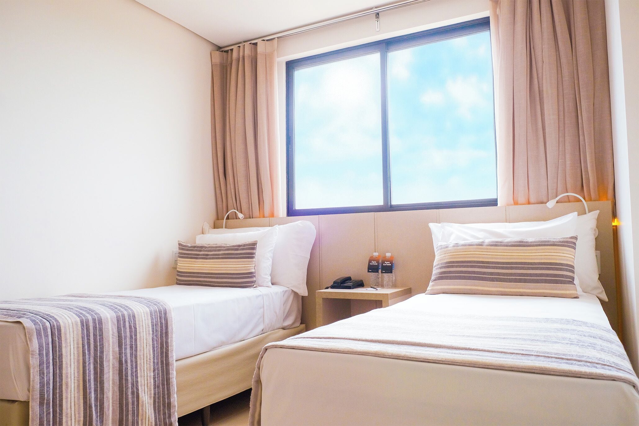 Hotellihuone, jossa on kaksi sänkyä, yöpöytä, jossa on puhelin ja pulloja, sekä ikkuna, jossa on verhot.