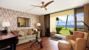 A modern living room with a sofa, armchair, and coffee table, a ceiling fan, and a large sliding glass door leading to a balcony with a view of the ocean.