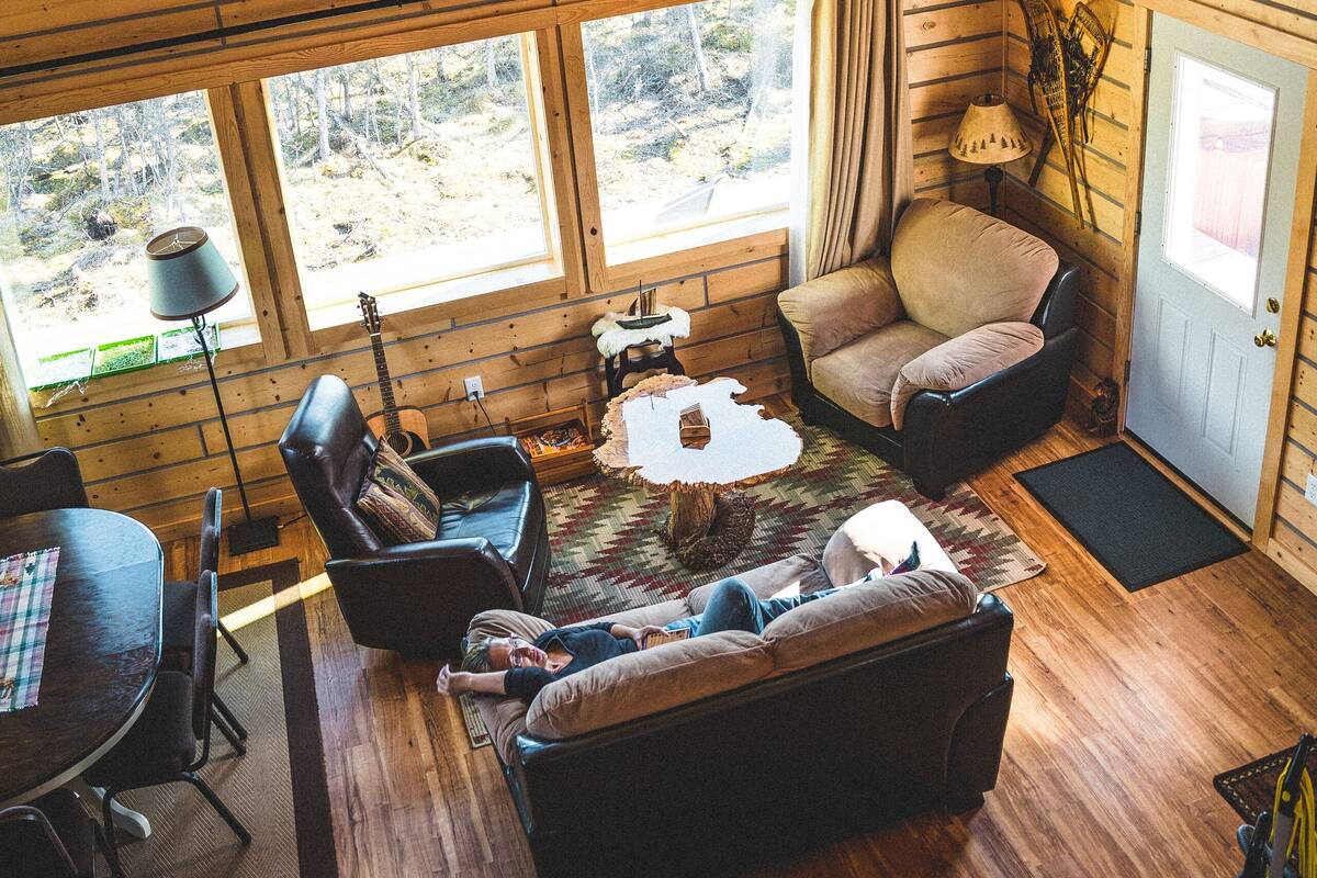 A cozy cabin interior with a high ceiling, wooden walls, a sofa, a dining table, and large windows offering a view of the outdoors.