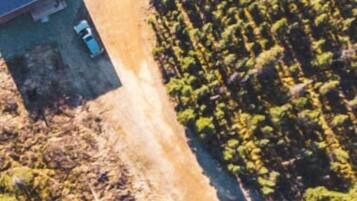 An aerial view of a rural area with a few buildings, a dirt road, and scattered vehicles.