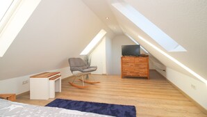 Une chambre moderne aménagée dans les combles, avec un plafond en pente, un plancher en bois, un fauteuil à bascule, un téléviseur posé sur un meuble en bois et une grande fenêtre laissant entrer la lumière naturelle.