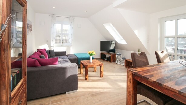 Un appartement-loft spacieux avec un plafond haut, un grand canapé d’angle gris, une table à manger en bois et un téléviseur dans l’espace de vie.