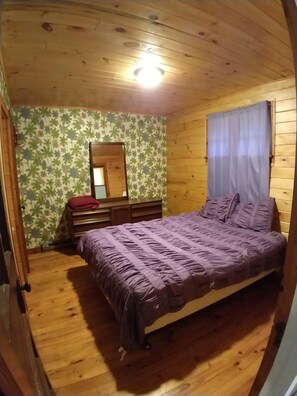Une chambre à coucher avec un plafond en bois, un lit recouvert d’une couverture violette, une commode, un miroir et une fenêtre avec un rideau.