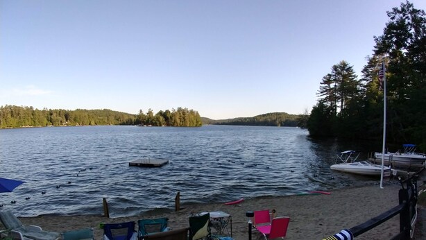 A lakeside view with a sandy beach, several colorful chairs, and a dock with boats. - #4 Adirondack FamilyFriendly Waterfront on LoonLake. Boating,fishing,swimming!  (Chestertown)