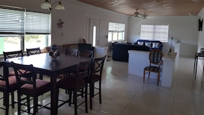 A spacious living area with a dining table, chairs, a sofa, and a small island with a chair. There are ceiling fans and a TV mounted on the wall. - Nature's Delight Lodge (Colonel Hill)