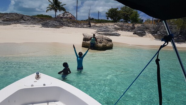 A boat with clear turquoise water, a sandy beach, and rocky terrain with palm trees in the background. - Nature's Delight Lodge (Colonel Hill)